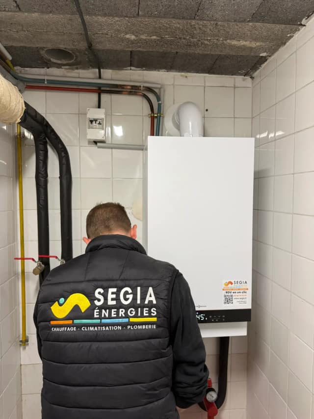 Technicien SEGIA de dos devant une chaudière murale Viessmann blanche posée dans un local carrelé, avec ses tuyaux de gaz et de chauffage